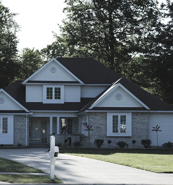 Suburban house with a dark roof and light blue siding, surrounded by green lawn and trees. A driveway leads to a white mailbox in front.