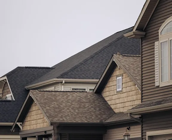 Roofs of suburban homes with varied peaks and angles under an overcast sky. The houses have brown siding and shingle roofing, evoking a cozy, calm atmosphere.