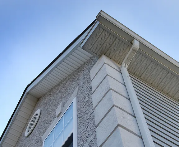 Low-angle view of a house corner showing a seamless gutter against a bright sky. The exterior walls have textured stucco and brick accents.